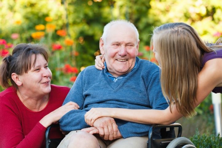 Pflegebedürftiger mann im Rollstuhl wird von rechts und links von pflegenden Angehörigen umsorgt.