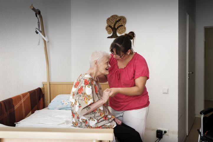Eine alte Frau sitzt auf einem Bett. Eine Pflegekraft hilft ihr aufzustehen. 