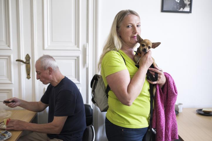 Ein Mann sitzt essend an einem Tisch und eine stehende Frau hält einen kleinen Hund auf dem Arm