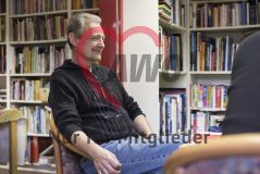 Ein Mann sitzt in einem Raum mit vielen Büchern an einem Tisch und hört zu und lächelt