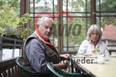 Ein älterer Mann und eine ältere Frau sitzen an einem Tisch und spielen Karten, er blickt dabei in die Kamera, sie auf ihr Blatt