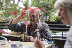 Ein älterer Mann sitzt an einem Tisch mit einer Frau, spielt Karten und kratzt sich dabei am Kopf