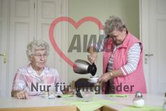 Eine ältere Frau sitzt am Tisch, während eine andere Frau ihr stehend Kaffee einschenkt