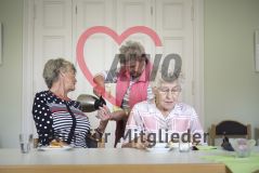Zwei ältere Damen sitzen am Tisch, während eine dritte hinter ihnen steht und Kaffee einschenkt