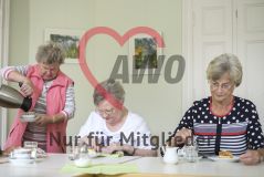 Zwei ältere Frauen sitzen am Tisch und essen Kuchen während eine dritte Frau neben ihnen steht und Kaffee einschenkt
