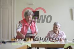 Eine ältere Frau sitzt am Tisch und sieht dabei zu, wie eine andere Frau stehend nach einem Stück Kuchen greift
