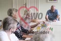 Eine Frau greift in eine Schüssel mit nummerierten Kugeln und spielt Bingo mit drei weiteren SeniorInnen, die mit am Tisch sitzen und auf die Spielfelder blicken