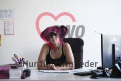 Eine Frau mit pinken Haaren, Piercings und auffälligem Schmuck sitzt auf einen Schreibtisch gestützt