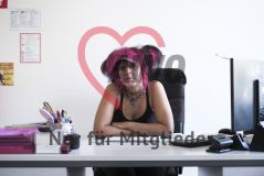 Eine Frau mit pinken Haaren, Piercings und auffälligem Schmuck sitzt auf einen Schreibtisch gestützt