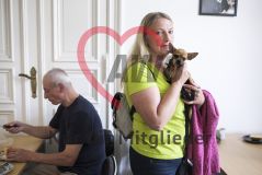 Eine Frau steht mit einem Chihuahua Hund auf dem Arm am Tisch, während ein Mann am Tisch sitzend einen Apfel aufschneidet