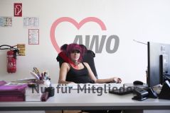 Eine Frau mit auffälliger pinker Frisur, Piercings und Halsschmuck sitzt an einem Schreibtisch 