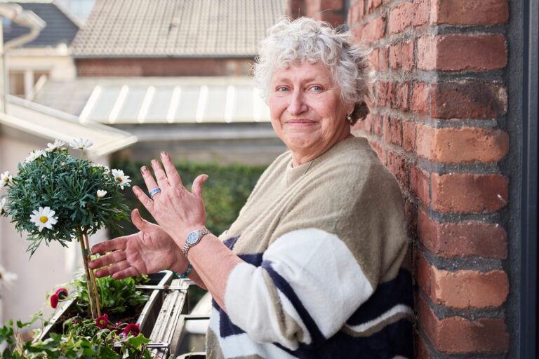Eine ältere Frau steht auf einem Balkon und pflanzt Blumen in einem Blumenkasten. Ihre Hände sind schmutzig von der Blumenerde.