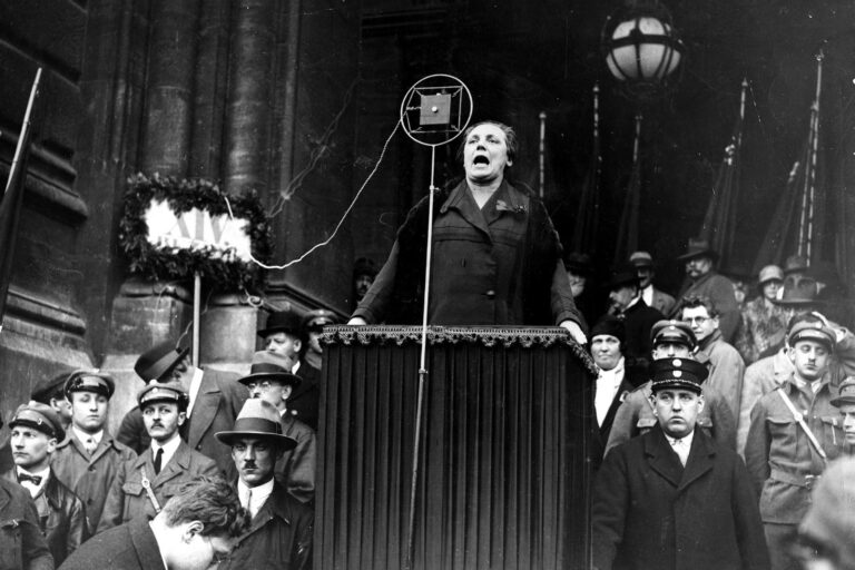 Schwarz-Weiß-Foto einer Frau, die an einem Rednerpult vor einer großen Menschenmenge spricht. Um sie herum stehen zahlreiche Personen.