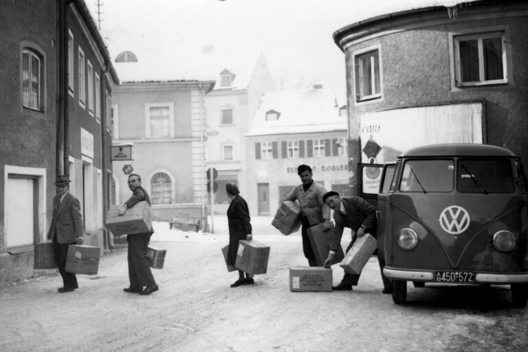 Winterliche Straßenszene in einer Stadt. Ein VW-Bus parkt am Straßenrand, während mehrere Männer und eine Frau Kartons und Pakete ausladen.
