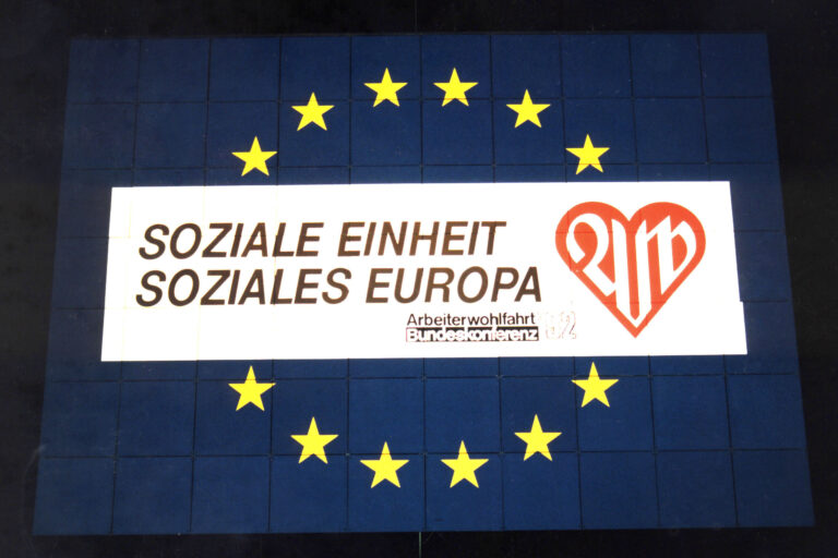 Plakat mit dem Slogan "Soziale Einheit, Soziales Europa" der Arbeiterwohlfahrt. Im Hintergrund ist die Europaflagge mit gelben Sternen auf blauem Grund zu sehen.