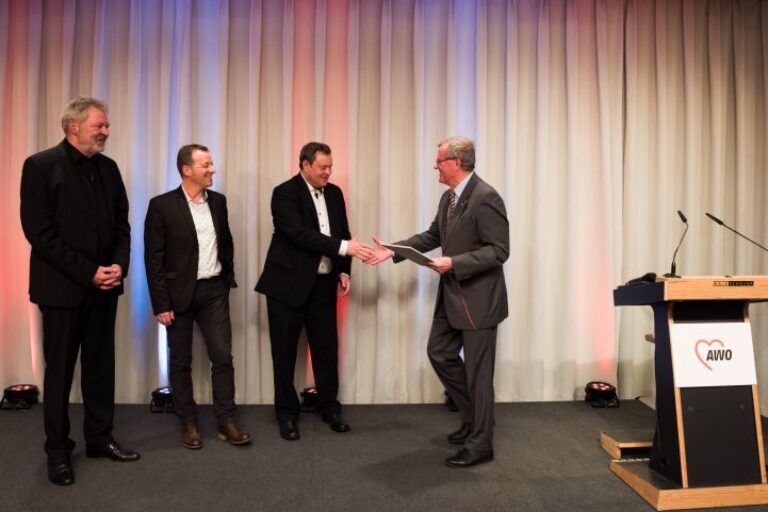 Vier Männer stehen auf einer Bühne vor einem hellen Vorhang. Einer der Männer schüttelt einem anderen die Hand und überreicht ihm eine Urkunde.