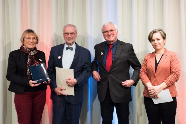 Vier Personen stehen vor einem hellen Vorhang und lächeln in die Kamera. Eine der Frauen hält eine dunkelblaue Mappe, einer der Männer hält eine weiße Urkunde.