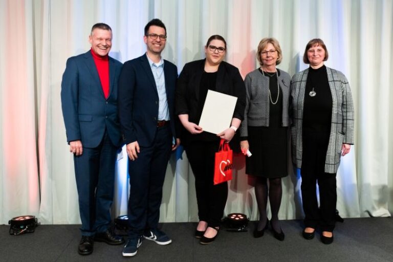 Fünf Personen stehen vor einem hellen Vorhang und lächeln in die Kamera. Eine der Frauen hält eine weiße Urkunde und eine rote AWO-Tasche in den Händen.