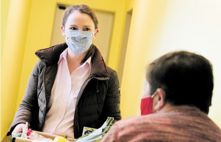 Eine Frau trägt eine Gesichtsmaske und hält eine Kiste mit Lebensmitteln. Sie steht in einem Flur und spricht mit einer Person, die ebenfalls eine Gesichtsmaske trägt.