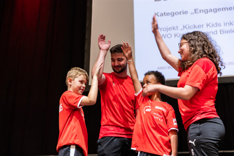 Zwei Kinder und zwei Erwachsene stehen auf einer Bühne und geben sich gegenseitig High-Fives. Sie tragen alle rote Sporttrikots mit dem AWO-Logo.
