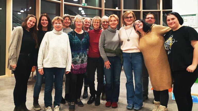 15 Personen stehen in einem Innenraum zusammen und lächeln in die Kamera. Die Gruppe besteht hauptsächlich aus Frauen unterschiedlichen Alters.