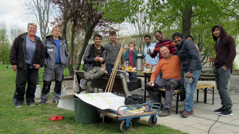 12 Personen stehen oder sitzen im Freien und lächeln in die Kamera. Alle tragen handwerklicher Arbeitskleidung. Sie befinden sich in einem Park.