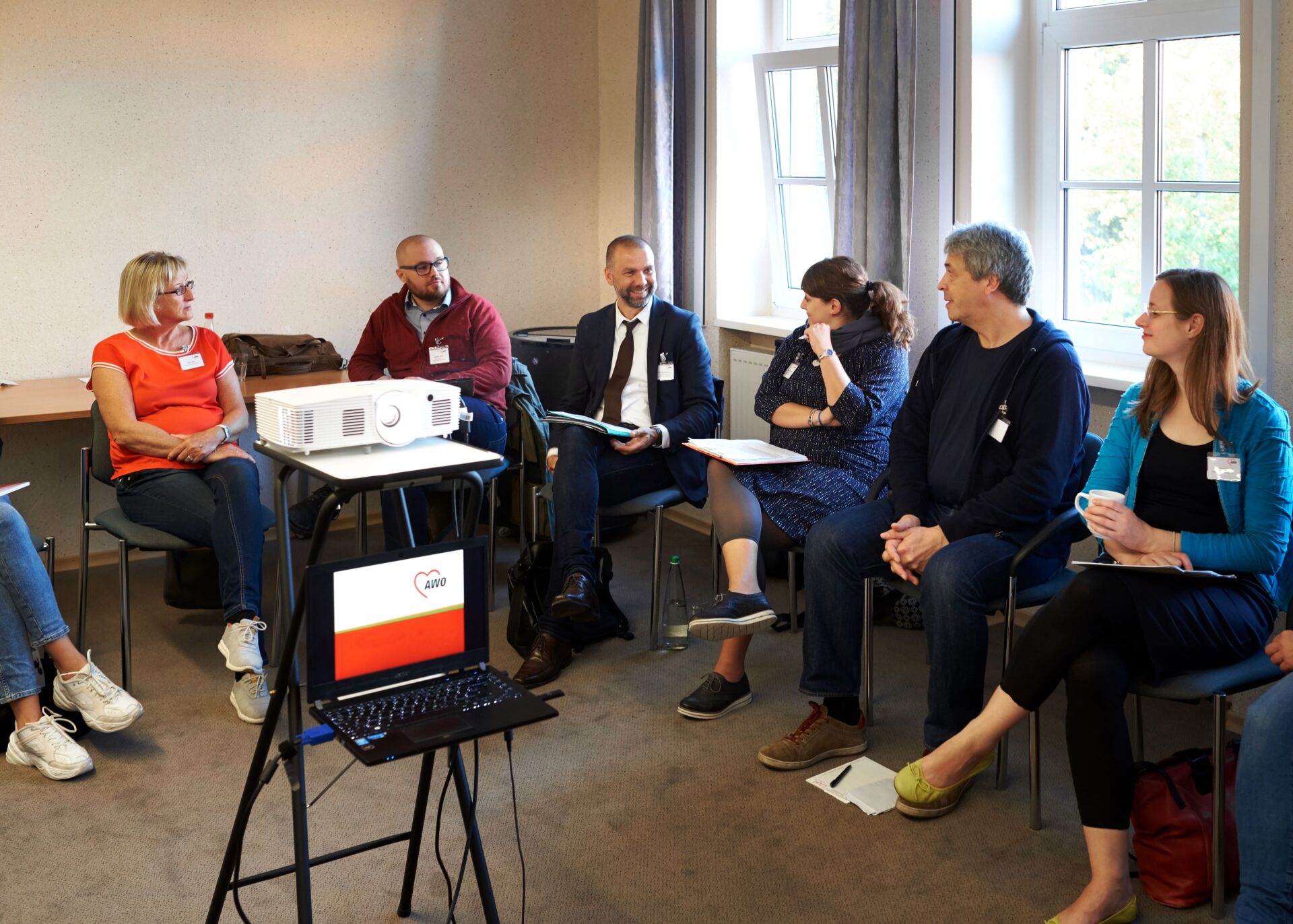 Eine Gruppe von Menschen sitzt in einem Seminarraum in einem Stuhlkreis und diskutiert. In der Mitte des Kreises steht ein kleiner Tisch mit einem Laptop und einem Projektor.