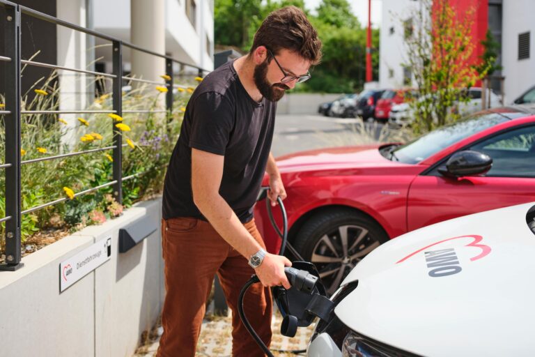 Ein Mann steht neben einem weißen Elektroauto mit dem AWO-Logo darauf und lädt es auf. Er hält das Ladekabel in der Hand und steckt es in das Auto.