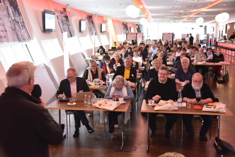Viele Personen sitzen an Tischen bei einer AWO-Versammlung. Ein Mann spricht in ein Mikrofon