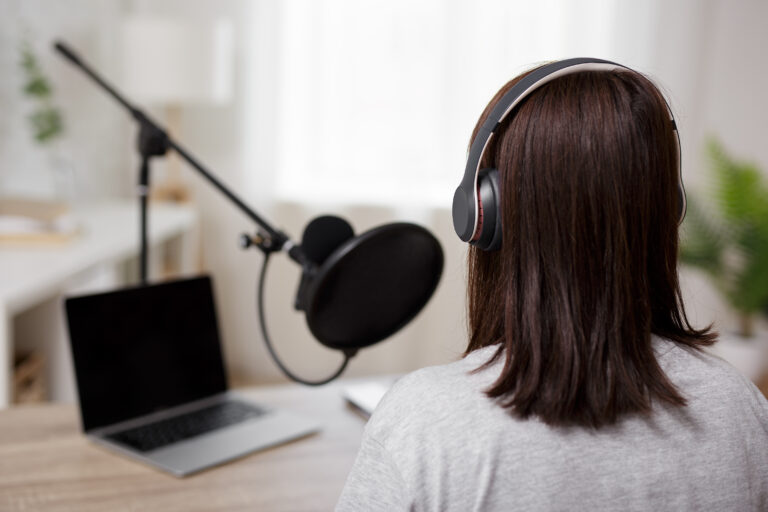 Rückansicht einer Frau mit halblangen braunen Haaren, grauem Shirt und Kopfhörern, die einen Audio-Podcast aufnimmt. Sie spricht in ein Mikrofon.