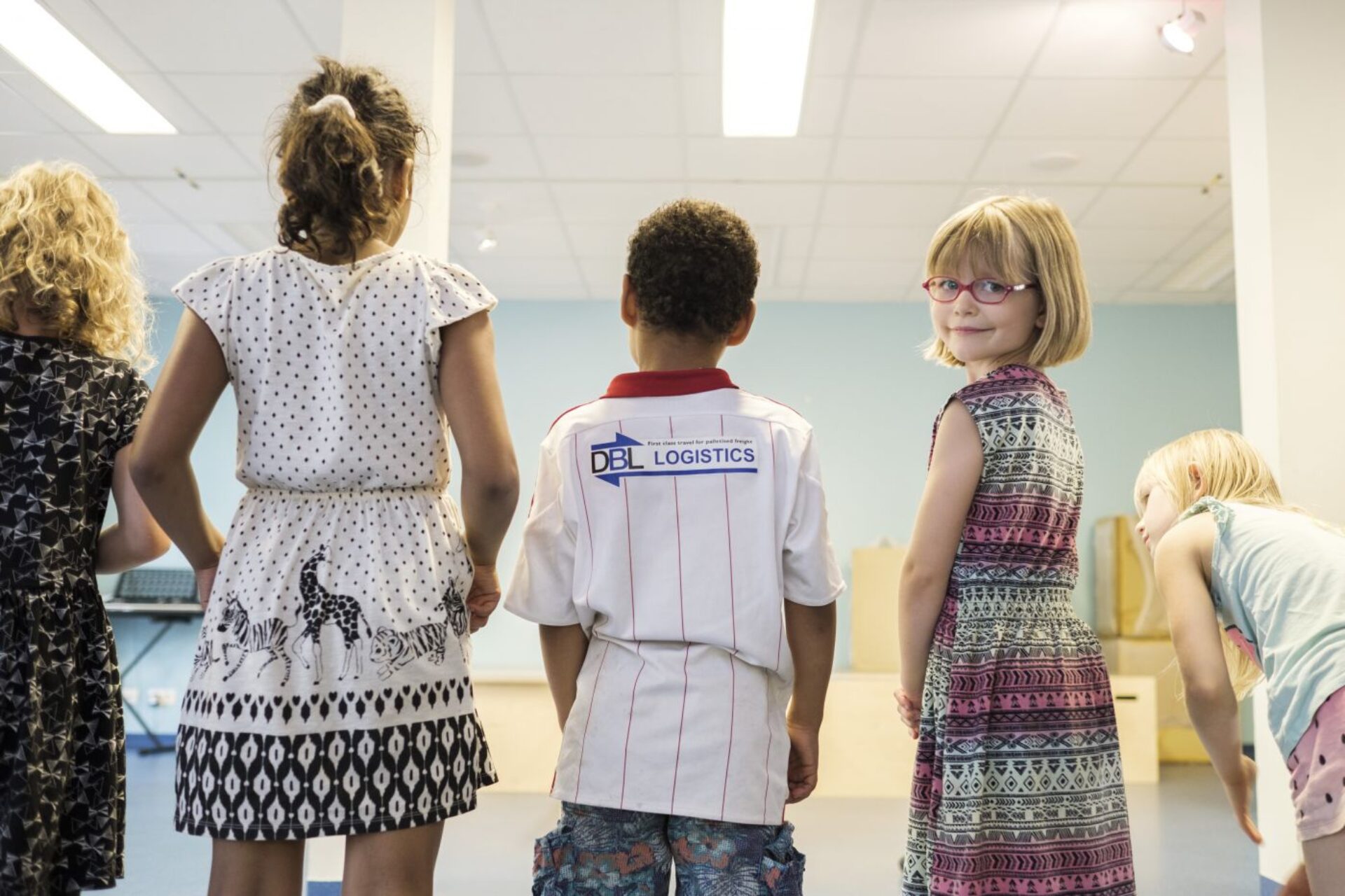 Mehrere Kinder stehen mit dem Rücken zur Kamera nebeneinander in einem Raum. Ein Mädchen mit Brille schaut lächelnd zurück.
