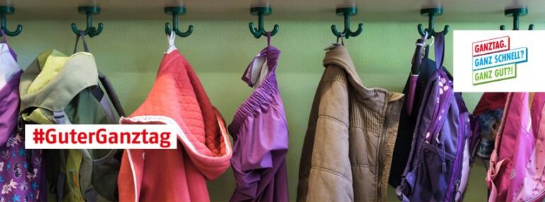 Jacken und Rucksäcke hängen an Haken in einer Garderobe. In einer Ecke steht: "#GuterGanztag"