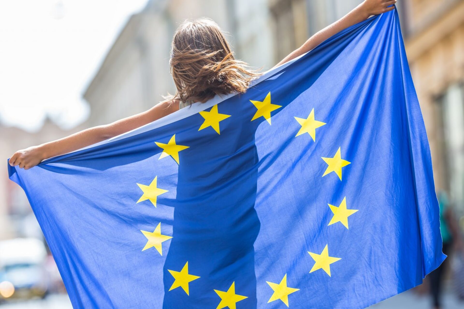 Junges Mädchen hält eine EU-Flagge mit ausgebreiteten Armen auf einer Straße.