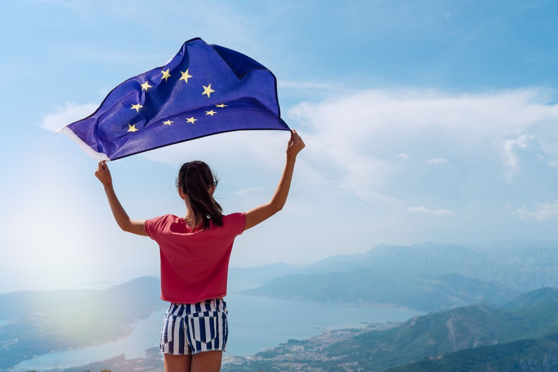 Mädchen steht auf einem Berg mit Blick aufs Meer und hält eine wehende EU-Flagge über dem Kopf.“