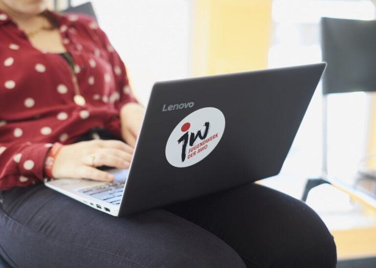 Eine Frau tippt auf einem Laptop auf ihrem Schoß. Auf dem Laptop klebt ein Sticker von jw - Jugendwerk der AWO.