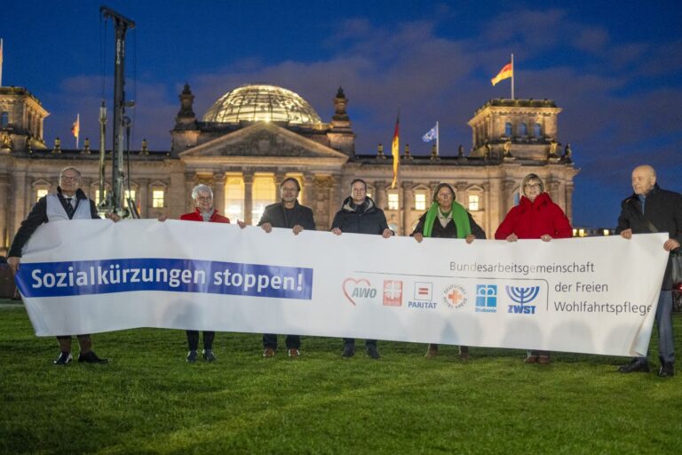 Eine Gruppe von Personen steht bei Nacht vor dem Bundestag. Sie halten ein großes Banner auf dem steht: "Sozialkürzungen stoppen!" steht.