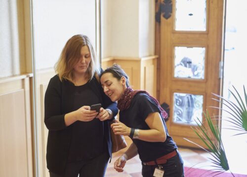 Zwei Frauen stehen in einem Eingangsbereich und schauen auf ein Smarthphone.