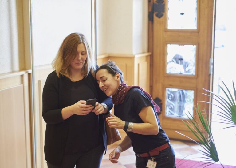 Zwei Frauen stehen in einem Eingangsbereich und schauen auf ein Smarthphone.