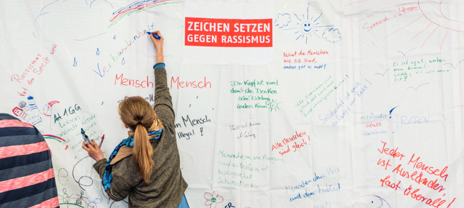 Eine Frau steht vor einer großen Wand, die mit verschiedenen Nachrichten und Zeichnungen gegen Rassismus bedeckt ist.