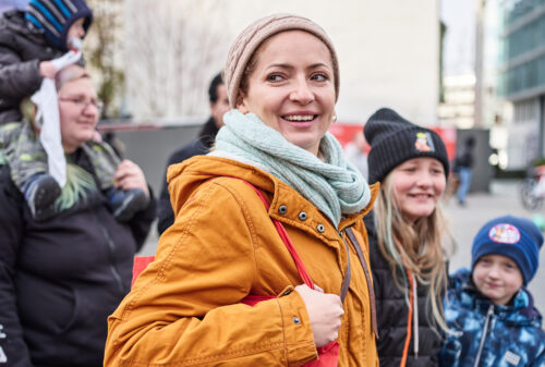 AWO-Mitarbeitende in ockerfarbener Jacke mit rotem AWO-Beutel, neben ihr eine Frau mit Kind auf den Schultern, ein lachendes Mädchen mit schwarzer Jacke und Mütze und ein Junge mit blauer Jacke und Mütze