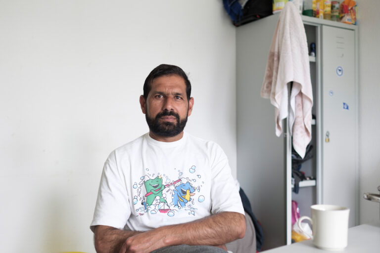 Ein Mann sitzt in einer Flüchtlingsunterkunft an einem Tisch, auf dem ein Kaffeebecher steht, neben ihm ein halb geöffneter Schrank.
