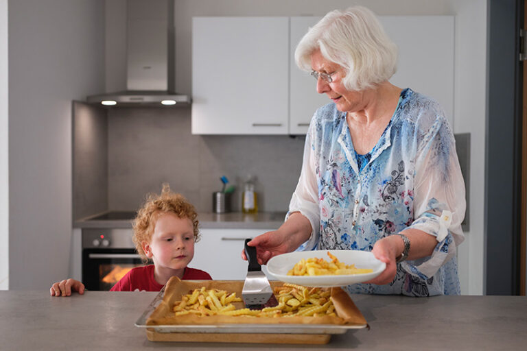 Eine Seniorin steht neben einem kleinen Jungen in der Küche und tut ihm Pommes Frites auf den Teller.