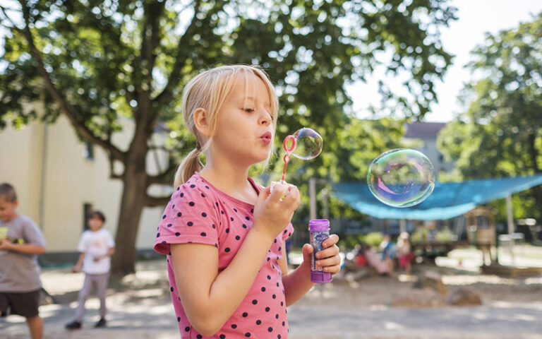 Ein Mädchen pustet Seifenblasen. Im Hintergrund spielen weitere Kinder.