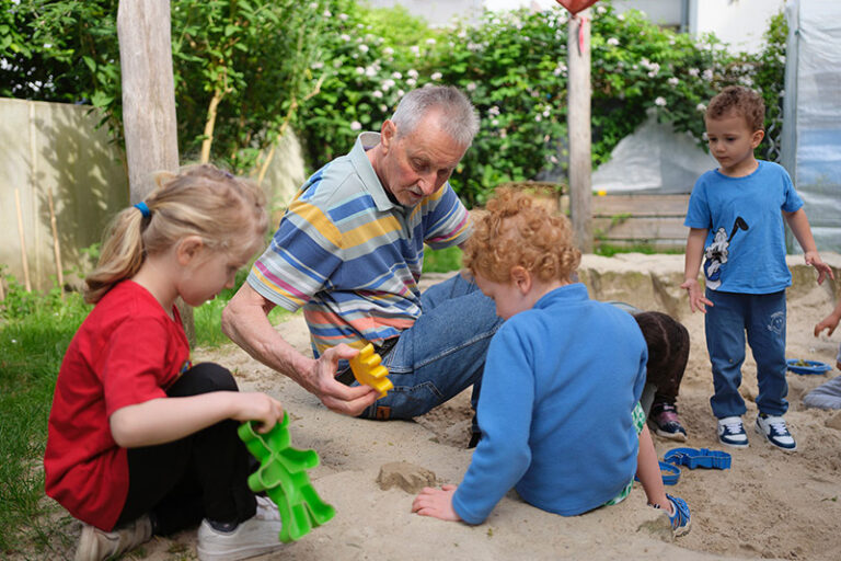Ein Senior sitzt mit Kindern im Garten in einer Buddelkiste.