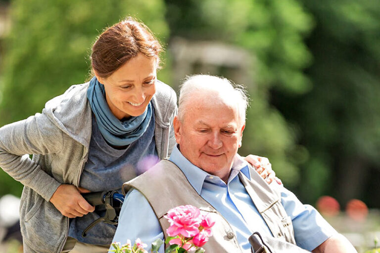 Ein Senior sitzt in einem Rollstuhl und wird draußen von einer jüngeren Frau geschoben.