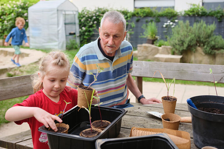 Ein kleines Mädchen und ein Senior setzen Jungpflanzen in eine Kiste. Im Hintergrund läuft ein Junge durch den Garten.