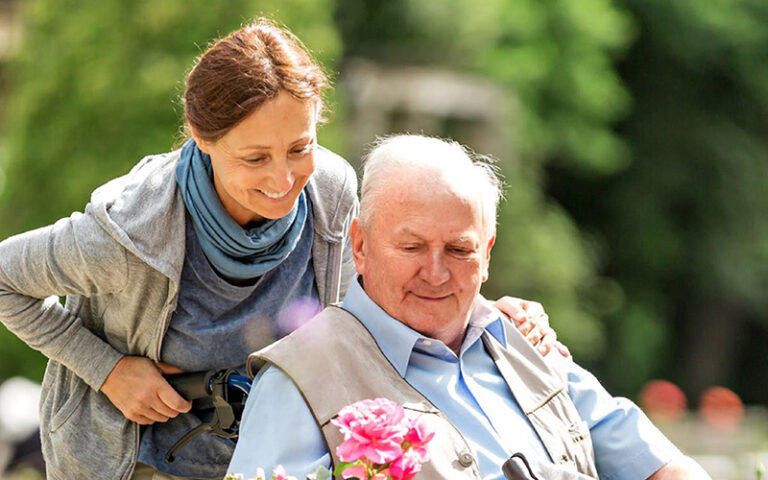 Eine lächelnde Frau schiebt einen Senior im Rollstuhl.