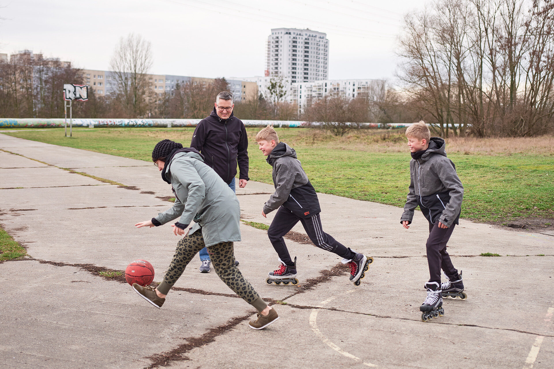 Mutter, Vater und zwei Söhne (beide auf Inlinern) spielen draußen Bastketball.