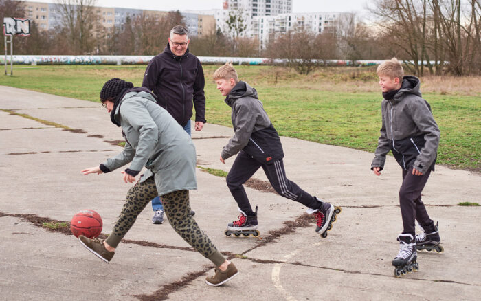 Mutter, Vater und zwei Söhne (beide auf Inlinern) spielen draußen Bastketball.
