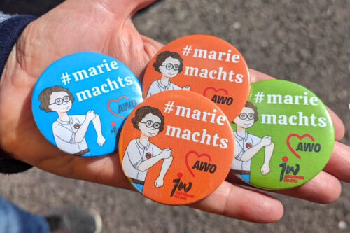 Vier Ansteck-Buttons - blau, grün, 2x orange - mit Marie macht's Logo liegen auf einer Hand.